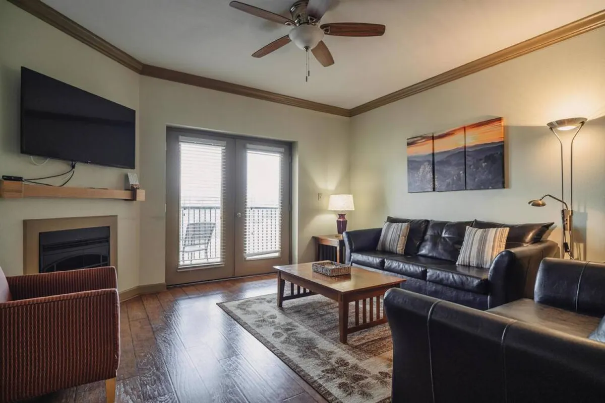 This image showcases a cozy living area featuring a comfortable sofa, a coffee table, tasteful artwork, and a fireplace. Natural light pours in, hinting at the presence of an outdoor balcony and giving the space a warm, inviting ambiance.