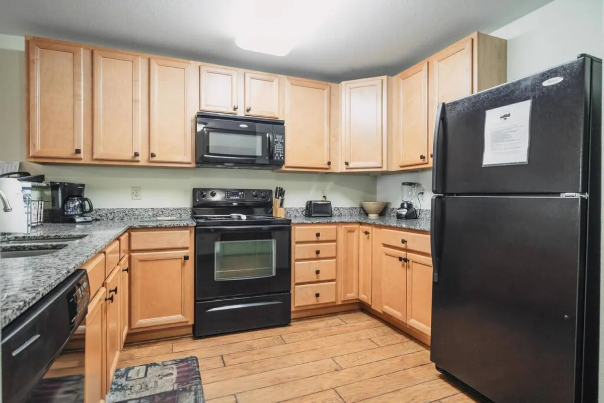 This image presents a kitchen featuring granite countertops, sleek black appliances including a stove and refrigerator, and warm wooden cabinets. The setup is spacious, inviting, and clearly suited for meal preparation and entertaining, with plenty of workspace and modern amenities visible.