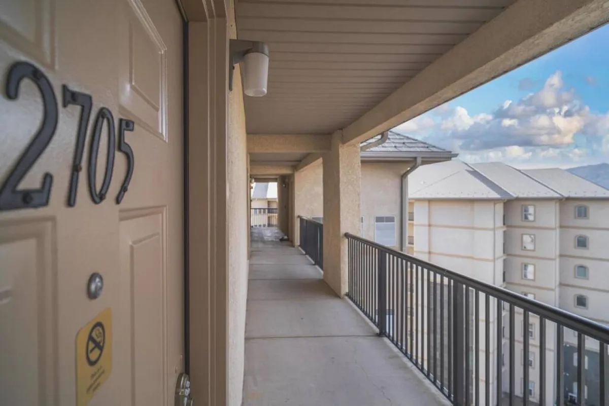 The exterior corridor with door number 2705 features a welcoming railing and light fixture. The space feels inviting, evoking the first moments of arrival and casual interactions with neighbors. Sunset lighting adds warmth to the scene.