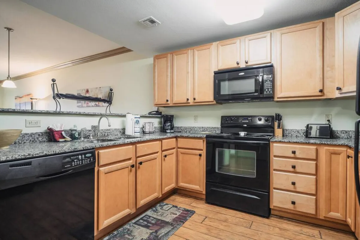 A welcoming kitchen featuring a spacious granite countertop, sleek black appliances, and ample wood cabinetry. The textured rug and open layout suggest ease of movement and accessibility for all guests. The space is bright, organized, and easy to navigate, suitable for guests with mobility considerations.