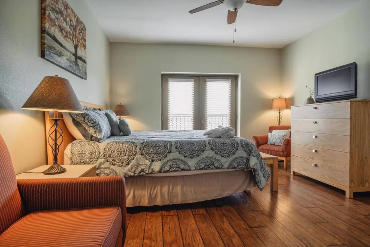 A comfortable bedroom scene with a bed, nightstand lamp, artwork on the wall, a dressing table, and a chair. The setup suggests a space for resting, reading, or watching TV, filled with natural daylight for a welcoming feel.