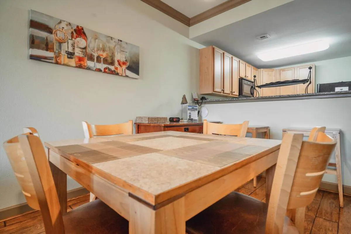 A cozy dining area with a wooden table and several chairs, complemented by visible kitchen cabinets and tasteful wall art. The arrangement offers generous space around the table, facilitating easy wheelchair access and movement. The scene feels inviting, inclusive, and practical for gatherings of all abilities.