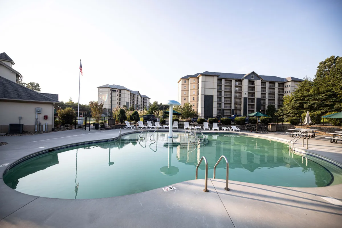 This image features an inviting outdoor swimming pool surrounded by lounge chairs, lush trees, and resort buildings in the background. Children can be seen playing in the water while adults relax nearby, creating a lively and family-friendly atmosphere.