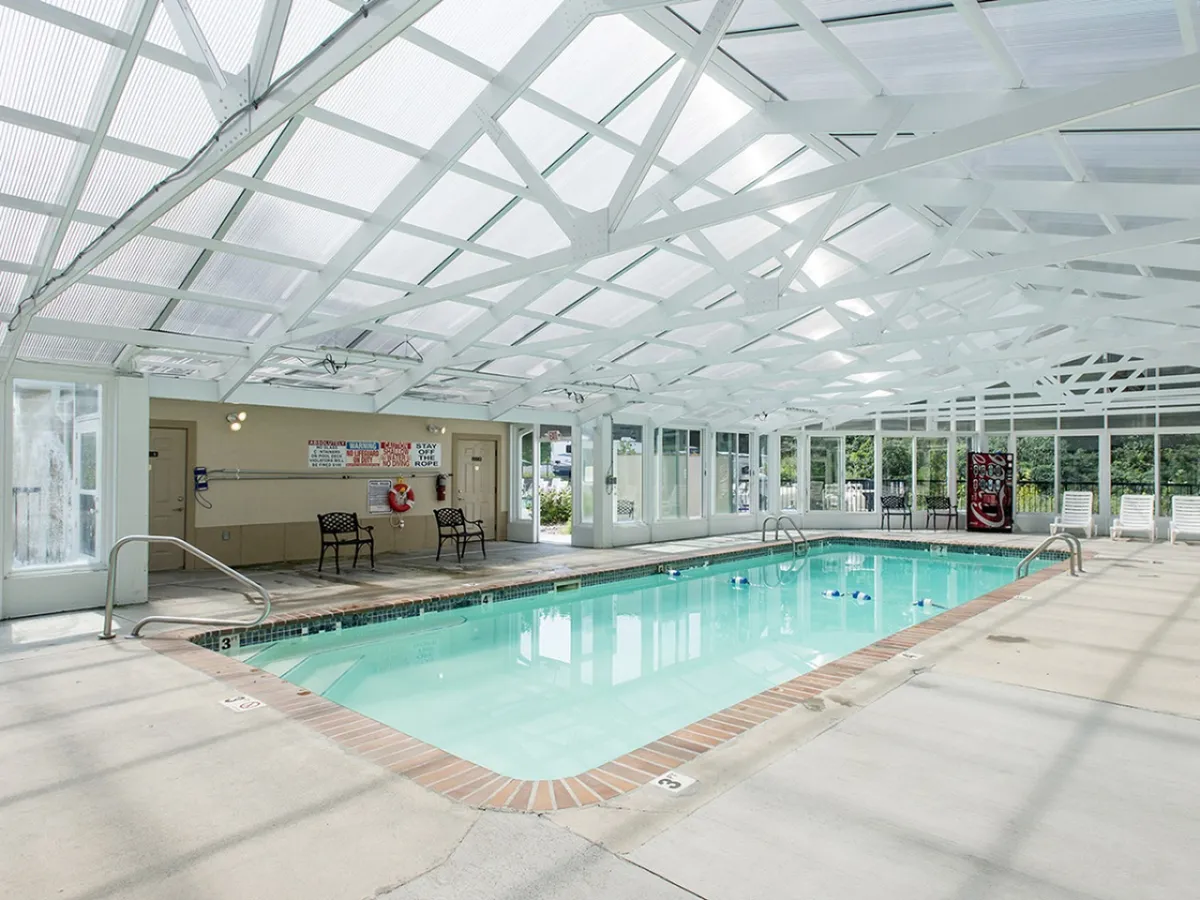 This image features the indoor swimming pool under a transparent roof, with lounge chairs lining the sides and safety equipment visible. The pool is filled with natural light, and the setup looks fresh and inviting, ideal for relaxing or swimming year-round.