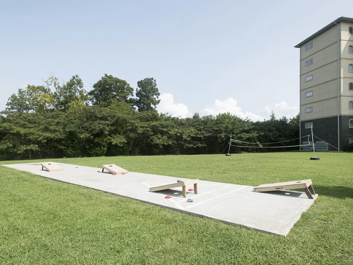 The scene shows a grassy lawn with cornhole boards set up, providing a vibrant space for outdoor games and family picnics. The resort building stands in the background, framing the communal recreation area.